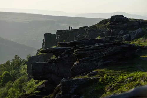 This landscape photograph captures Curbar Edge in the Peak District, located in Derbyshire, United Kingdom, during a spring evening. The main subject of the image is the prominent rocky escarpment of Curbar Edge, which rises above the surrounding rural terrain and showcases the dramatic geological formations typical of the Peak District. In the distance, two figures stand atop the rocks, emphasizing the scale and natural beauty of the area. The scene is bathed in soft evening light, casting long shadows across the rugged landscape and highlighting the lush greenery of spring nature below. The rolling hills and layered cliffs demonstrate the unique character of this rural region in the United Kingdom.